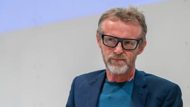 Ein Mann mittleren Alters mit graumeliertem Bart, kurzen schütteren Haaren und einer kantigen schwarzen Kunststoffbrille.