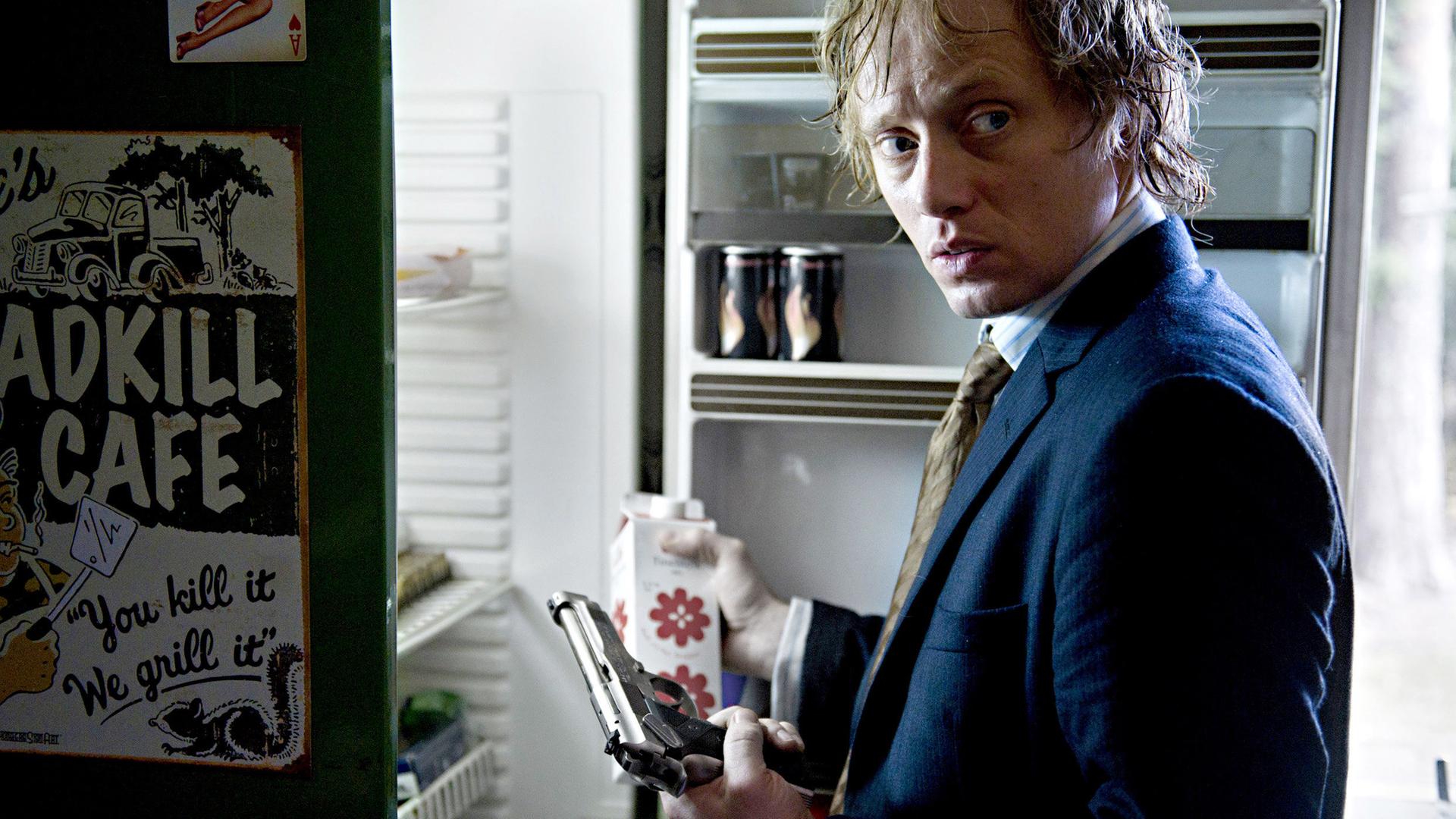 Headhunter Roger Brown (Aksel Hennie) steht mit Pistole bewaffnet vorm Kühlschrank.