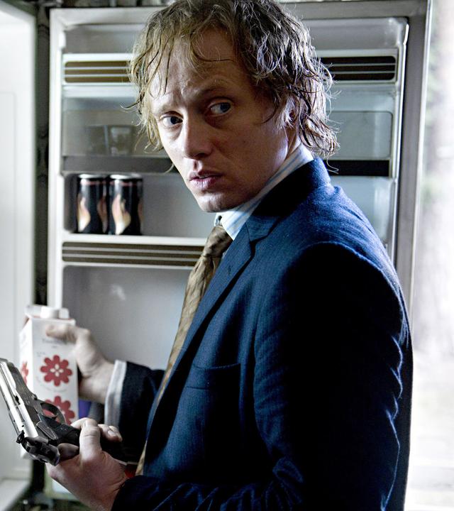 Headhunter Roger Brown (Aksel Hennie) steht mit Pistole bewaffnet vorm Kühlschrank.