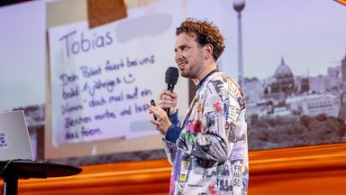 Joab Nist steht auf der rechten Seite des Bildes. Auf der linken Bildseite steht ein Laptop auf einem Stehtisch. Im Hintergrund ist ein Bild der Berliner Skyline zu sehen. Er trägt einen bunten Pullover und hält das Mikrofon in seiner rechten Hand.