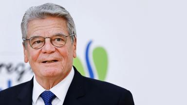 Ein älterer Mann mit kurzen grauen Haaren und Brille in einem dunklen Anzug mit blauer Krawatte.