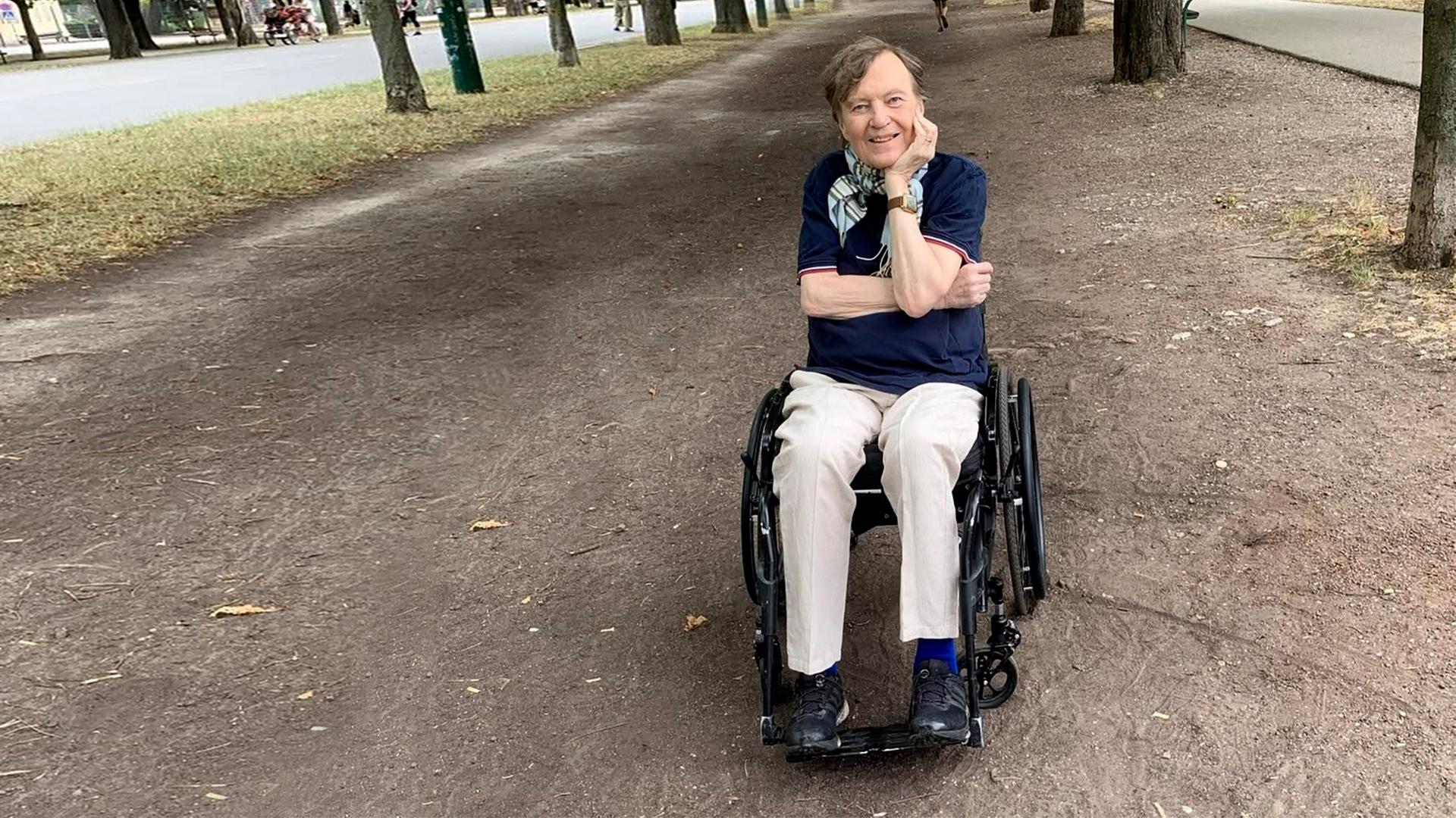 Mann sitzt im Rollstuhl auf einer Allee und lächelt.