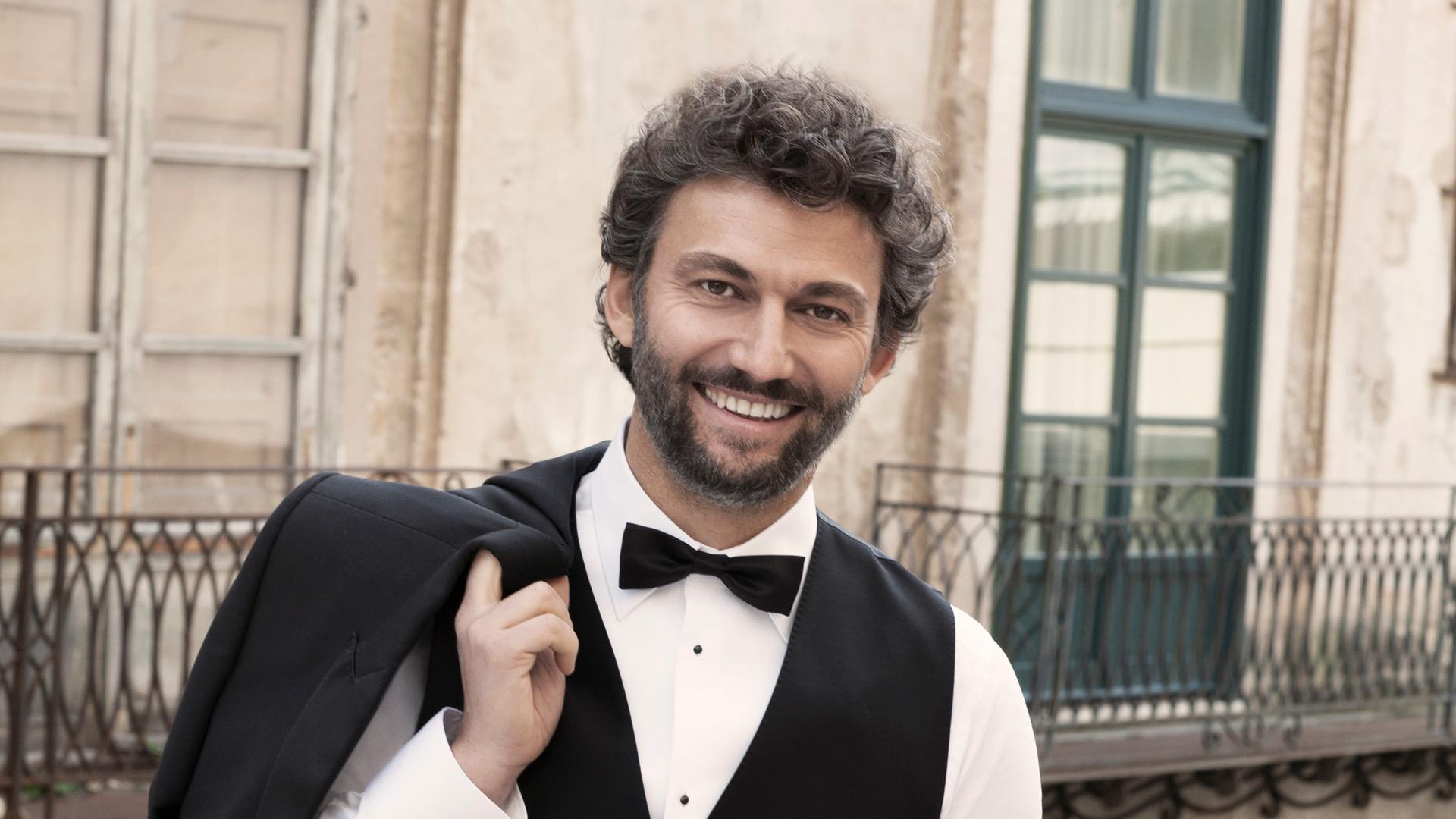 Jonas Kaufmann steht auf einer Brücke und lächelt. Er trägt einen dunklen Anzug und ein weißes Hemd. Sein Jacket hat er lässig über die rechte Schulter gelegt und hält es mit seiner rechten Hand fest.