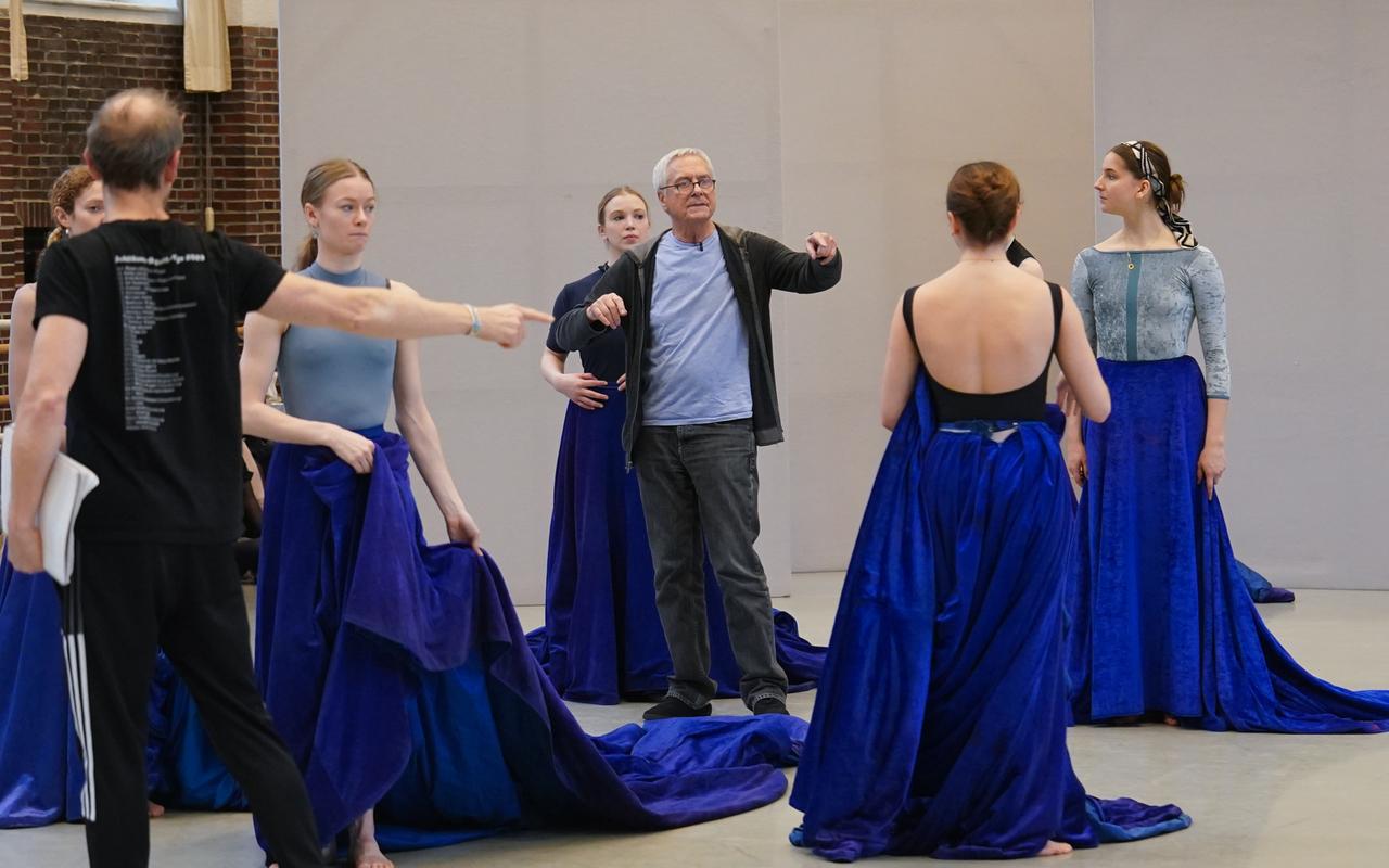 Zwei Männer stehen zwischen Tänzerinnen in langen blauen Röcken und geben ihnen gestikulierend Anweisungen.