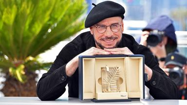 Der Schauspieler John Travolta, ein schwarz gekleideter Mann mit Baskenmütze, brille und Bart stützt sich lächelnd auf der goldenen Ehrenpalme von Cannes ab, einer goldenen Trophäe in einer beige ausgeschlagenen Schatulle.