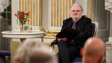 Ein mittelalter Mann mit zu einem Zopf zusammengebundenen grauen Haaren und grauem Bart sitzt auf einem Stuhl an einem runden Tisch und hält eine aufgeschlagene Kladde in der Hand.