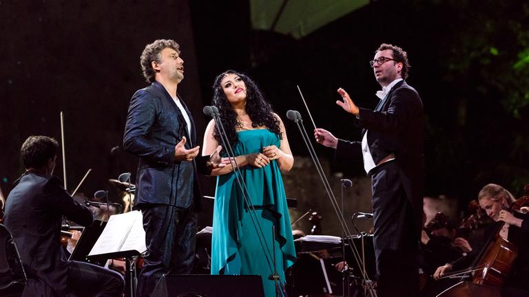 Jonas Kaufmann und Anita Rachvelishvili stehen vor dem Orchster auf der Bühne. Jonas Kaufmann singt. Am rechten Bildrand steht dirigierend Jochen Rieder.