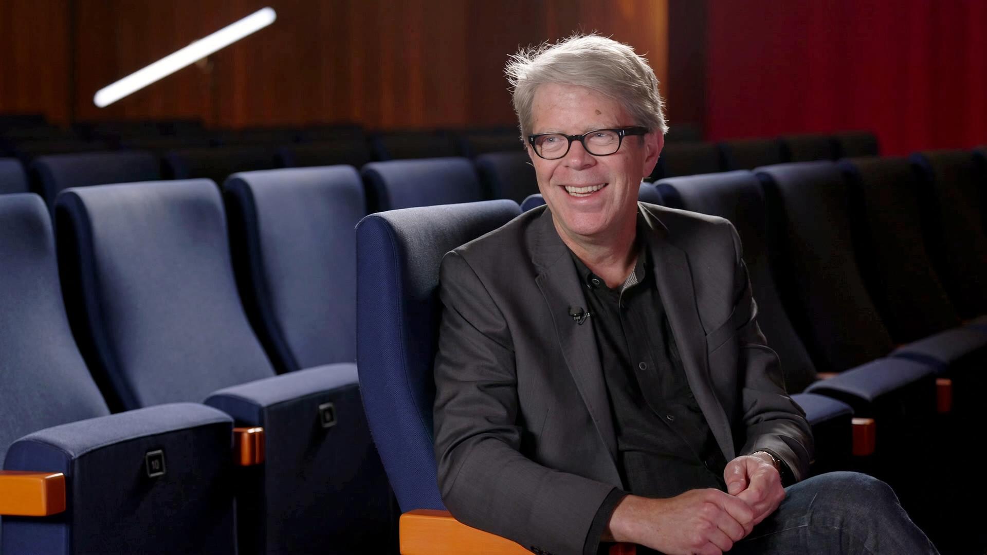 Jonathan Franzen sitzt lächelnd in einem leeren Kinosaal auf einem blauen Sessel mit orangefarbenen Armlehnen, bekleidet mit dunklem Sakko und Brille.