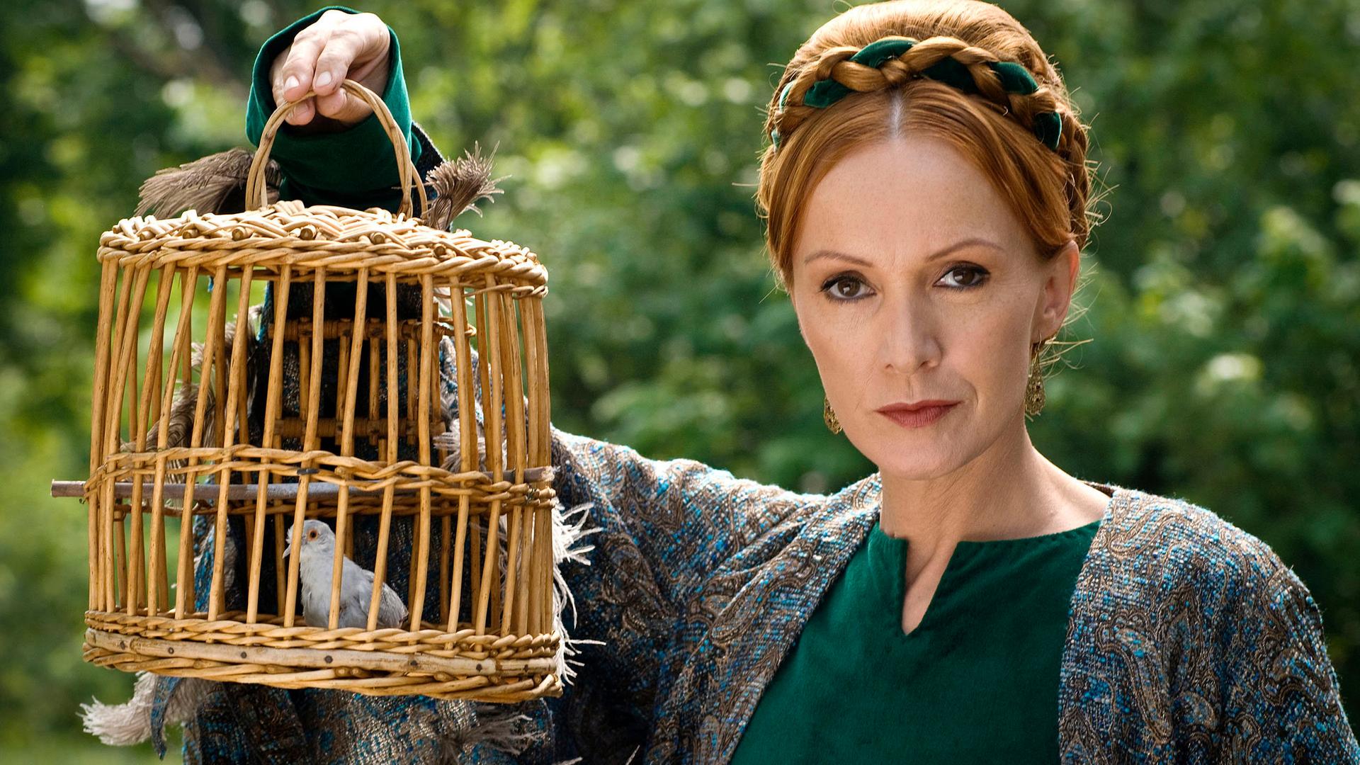 Die Zauberin (Katja Flint) hält einen Weidenkäfig mit einem kleinen weißen Vögelchen hoch und blickt boshaft.