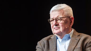 Porträtfoto eines älteren Mannes mit grauen Haaren, Brille und braunem Tweed-Anzug.