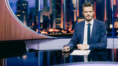 Florian Schroeder sitzt am Schreibtisch im Studio seiner neuen Show "JOURNAL DELUXE". Im Hintergrund ist durch ein ovales Fenster eine Skyline mit Hochhäusern zu sehen.