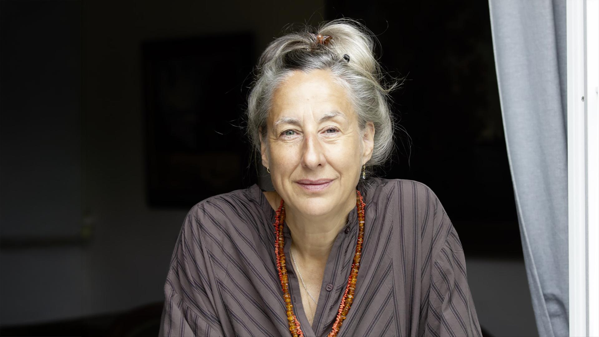 Porträtfoto einer Frau mittleren Alters mit hochgesteckten graublonden Haaren, die eine lange doppelreihige orangefarbene Edelsteinkette trägt.