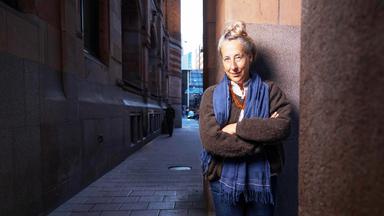 Eine Frau mittleren Alters mit hochgesteckten graublonden Haaren und bekleidet mit einem Strickpullover und Schal steht an einer Straßenecke und lächelt in die Kamera.