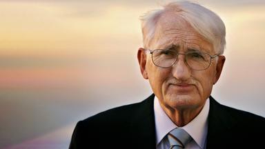 Porträtfoto eines älteren Mannes mit kurzen weißen Haaren und Schnurrbärtchen, der eine Brille und einen Anzug trägt und vor einem Sonnenuntergang steht.