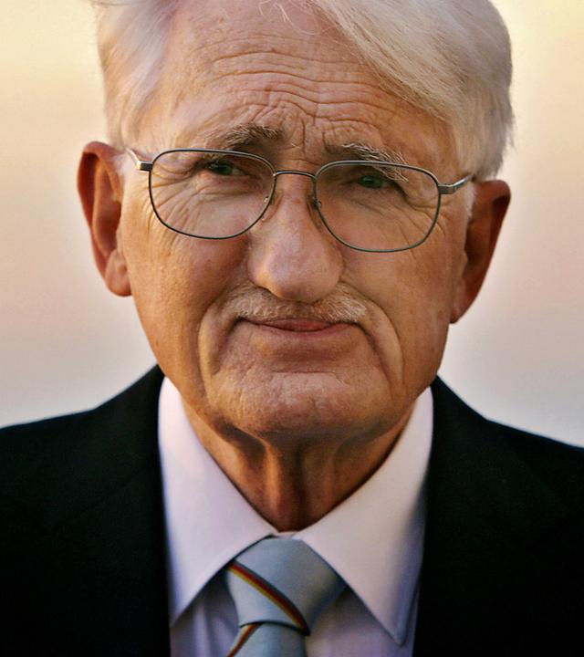 Porträtfoto eines älteren Mannes mit kurzen weißen Haaren und Schnurrbärtchen, der eine Brille und einen Anzug trägt und vor einem Sonnenuntergang steht.