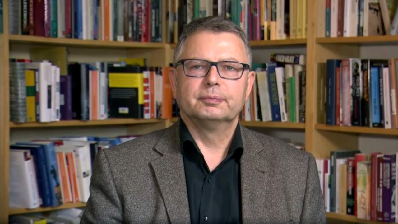 Ein Mann mit Brille und grauem Haar steht vor einem Bücherregal