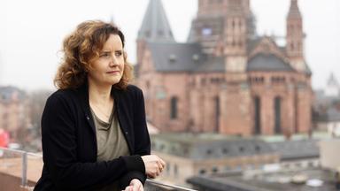 Eine Frau mit lockigen mittelblonden Haaren steht auf einem Balkon und schaut in die Ferne. Hinter ihr ist der Mainzer Dom zu sehen.