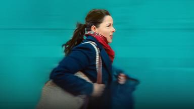 Julie (Laure Calamy), eine junge Frau mit Pferdeschwanz, in blauem Mantel, rotem Halstuch und mit einer Tasche um die Schulter, eilt gehetzt voran. Im Hintergrund eine grüne Wand oder Fassade. Wegen der Geschwindigkeit ist das Bild leicht unscharf.