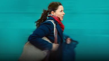 Julie (Laure Calamy), eine junge Frau mit Pferdeschwanz, in blauem Mantel, rotem Halstuch und mit einer Tasche um die Schulter, eilt gehetzt voran. Im Hintergrund eine grüne Wand oder Fassade. Wegen der Geschwindigkeit ist das Bild leicht unscharf.