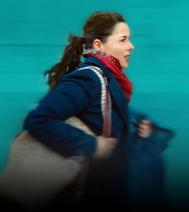 Julie (Laure Calamy), eine junge Frau mit Pferdeschwanz, in blauem Mantel, rotem Halstuch und mit einer Tasche um die Schulter, eilt gehetzt voran. Im Hintergrund eine grüne Wand oder Fassade. Wegen der Geschwindigkeit ist das Bild leicht unscharf.