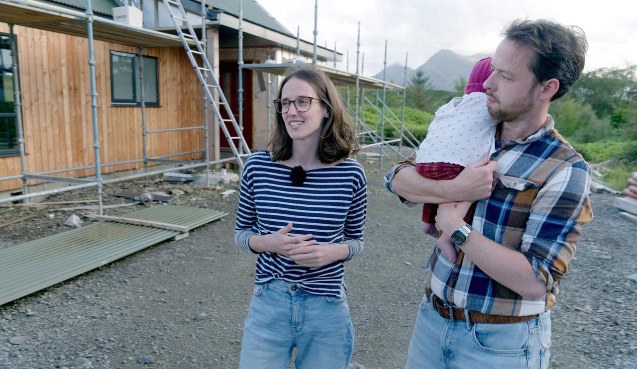 Das Bild zeigt eine Szene im Freien vor einem unvollendeten Gebäude. Im Vordergrund stehen zwei Personen: eine Frau und ein Mann. Die Frau trägt ein gestreiftes Oberteil und hat schulterlanges Haar. Sie spricht und wirkt freundlich. Der Mann hält ein kleines Kind in den Armen, das eine weiße Kleidung mit Punkten trägt. Er ist in einem karierten Hemd gekleidet und schaut ernst.   Im Hintergrund sind Gerüste und Baustellenmaterialien zu sehen, was darauf hindeutet, dass sich das Gebäude im Bau befindet. Der Himmel ist teils bewölkt, und im Hintergrund sind einige Hügel oder Berge zu erkennen. Der Boden besteht aus Kies und Erde, was typisch für einen Bauplatz ist.