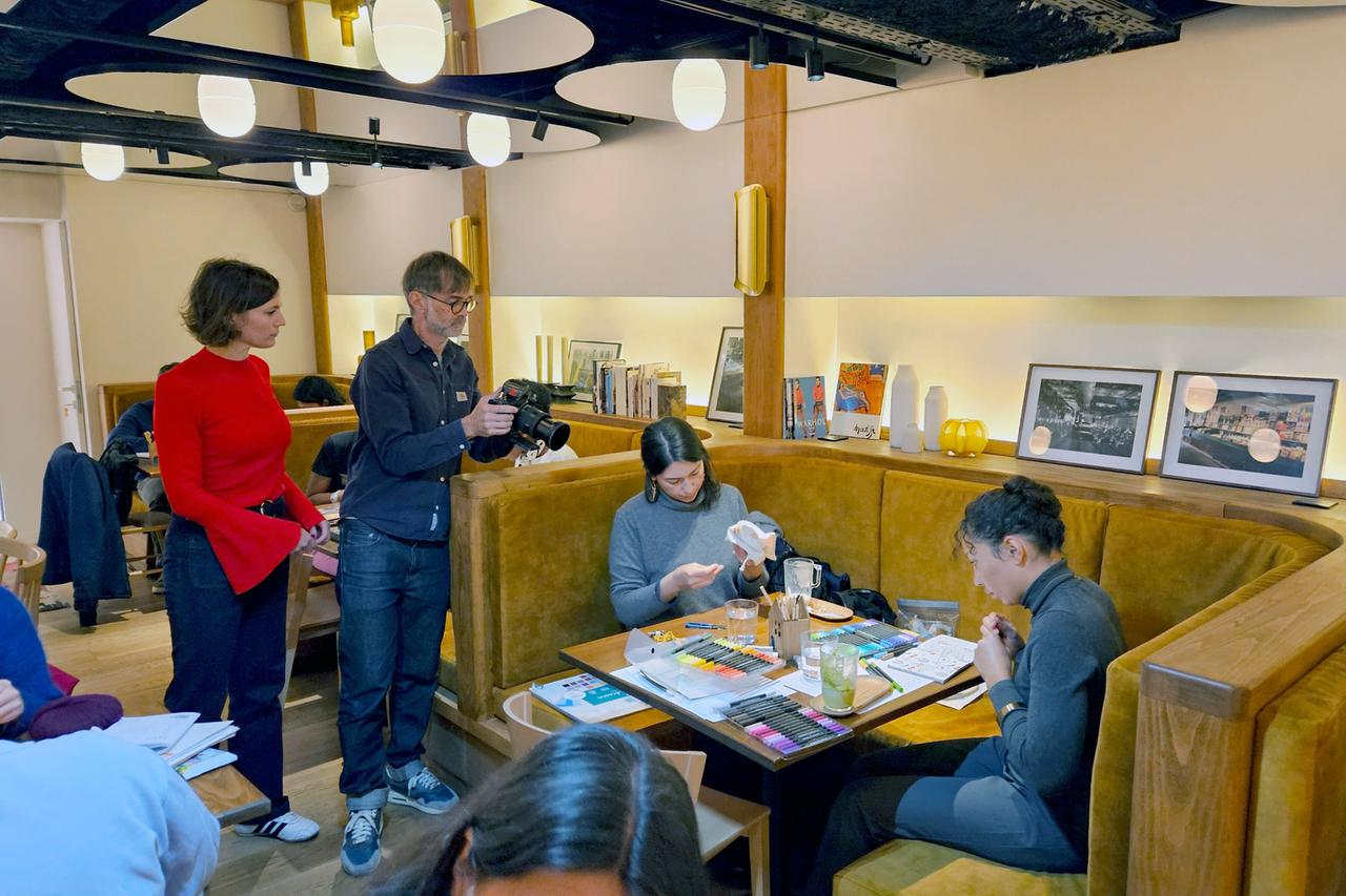 Stefania Tsakiraki steht mit zwei jungen Erwachsenen in einem hellen Raum mit großen Fenstern und Pflanzen. Sie sprechen miteinander, die Smartphones liegen außer Sichtweite.