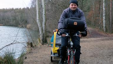 Björn ist mittig im Bild auf seinem Fahrrad zu sehen. Er trägt eine schwarze Mütze und eine graue Jacke. Rechts von ihm sieht man das Ufer eines Sees. Links von ihm führt ein Weg in einen Wald.