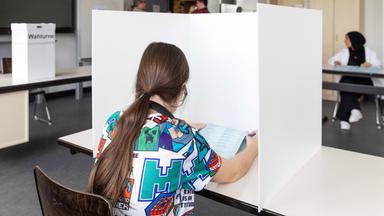Rückansicht einer jungen Frau beim Ausfüllen eines Wahlzettels, in einem Klassenzimmer