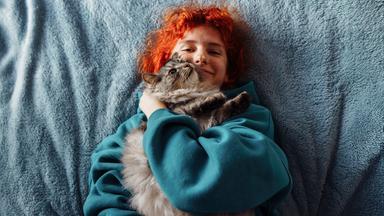 Eine junge Frau mit roten Haaren liegt auf dem Bett und hat eine grau getiegerte Katze im Arm auf sich lieben.