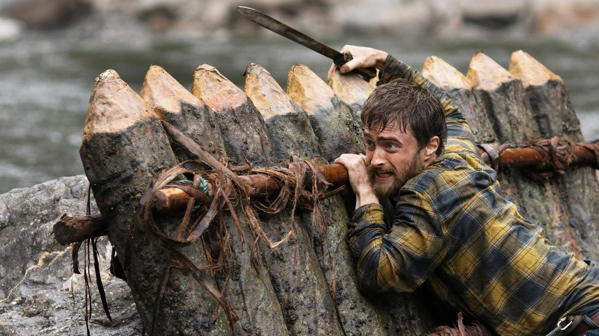 Yossi (Daniel Radcliffe) klammert mit Machete an schräg an Felsen aufliegendem Floß.