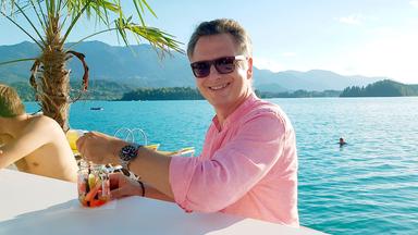 Moderator Michael Friemel sitzt an der Strandbar mit Palmen am Faaker See. Hinter ihm türkises Wasser