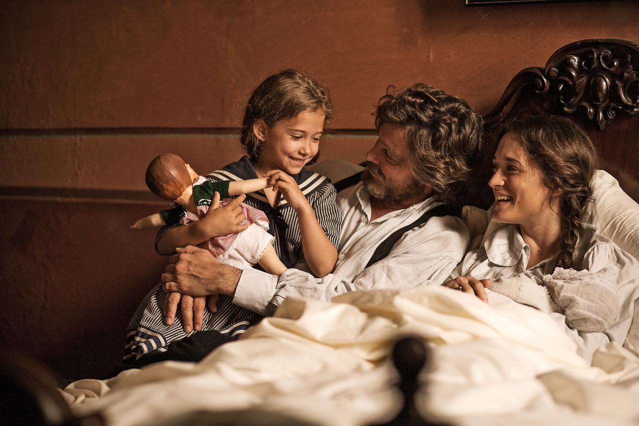 Das Bild zeigt eine Szenerie im Innenraum, möglicherweise in einem Schlafzimmer oder einer ähnlichen gemütlichen Umgebung.   Im Vordergrund sind drei Personen zu sehen, die auf einem Bett sitzen. Eine Frau und ein Mann lächeln sich an, während sich ein kleines Mädchen zwischen ihnen befindet. Das Mädchen hält eine Puppe in der Hand, die eine traditionelle Kleidung trägt.   Die Frau hat lange, braune Haare, die offen oder leicht gewellt sind, und trägt ein einfaches, helles Oberteil. Der Mann hat einen Bart und trägt ein Hemd mit einem Hosenanzug. Beide Erwachsenen scheinen erfreut und entspannt zu sein.   Die Wände des Raumes sind in warmen Erdtönen gehalten, und das Bett hat ein schmiedeeisernes Kopfteil. Eine Decke aus hellem Stoff verdeckt den unteren Teil des Bettes. In der Umgebung sind keine spezifischen Details zu erkennen, die auf den genauen Ort oder die Zeitperiode hinweisen.   Die Szene strahlt eine familiäre und herzliche Atmosphäre aus.