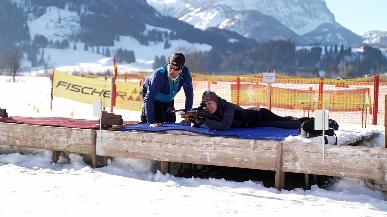 Das Bild zeigt eine winterliche Szene im Kaisergebirge, umgeben von schneebedeckten Bergen. Im Vordergrund sieht man eine Biathlon-Schießstand, auf dem eine Frau auf einer Matte liegt und mit einem Biathlon-Gewehr zielt. Sie trägt eine sportliche Kleidung, die aus einer schwarzen Weste und einer Mütze besteht. Neben ihr steht ein Mann, der sie anleitet. Er trägt eine blaue Jacke und Schützer auf den Augen. Im Hintergrund sind schneebedeckte Hügel und einige Bäume sowie ein abgesperrter Bereich sichtbar, der mit einem gelben Banner mit dem Schriftzug „FISCHER“ gesäumt ist. Der gesamte Bereich ist von Schnee bedeckt, was die winterliche Atmosphäre unterstreicht.