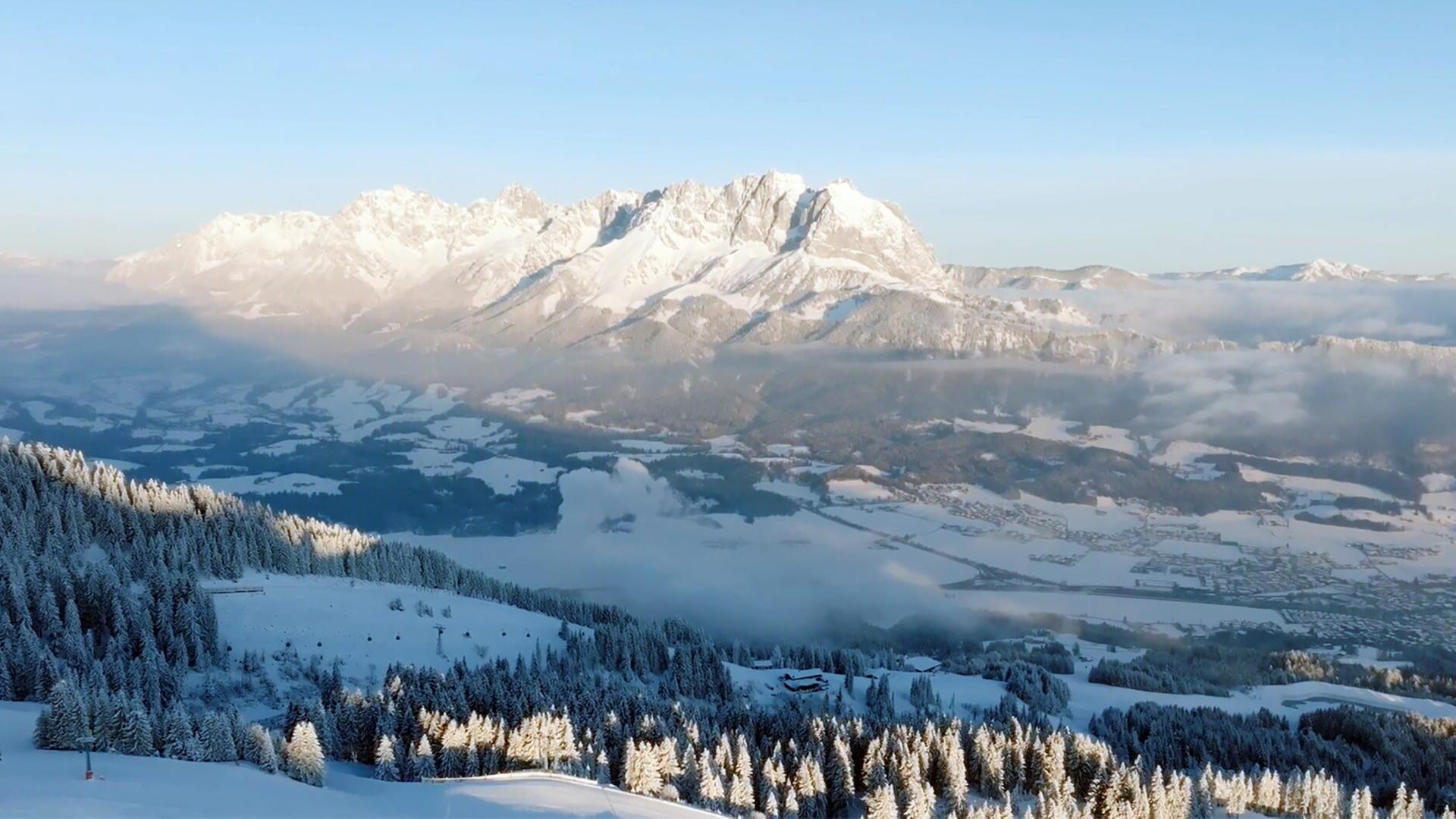 Das Bild zeigt eine beeindruckende winterliche Landschaft im Kaisergebirge in Tirol. Die Szenerie ist geprägt von hohen, schneebedeckten Bergen, die im Hintergrund thronen, während im Vordergrund dichte Nadelwälder zu sehen sind, die ebenfalls mit Schnee bedeckt sind. Die Bäume sind teilweise sonnendurchflutet und erscheinen heller, während der Rest der Landschaft schattiger ist.   Der Himmel ist klar und blau, was die Kälte und Frische des Morgens unterstreicht. Im Tal erstreckt sich eine weite, schneebedeckte Landschaft, die von einzelnen Nebelschwaden durchzogen ist. Hier sind auch sporadisch Wohngebäude und Straßen sichtbar, die sich durch die winterliche Landschaft schlängeln.   Insgesamt vermittelt das Bild eine ruhige und friedliche Winteratmosphäre, die die natürliche Schönheit der Umgebung hervorhebt. Es handelt sich um eine Szene, die typisch für die Region St. Johann in Tirol ist und die sportlichen Abenteuer sowie das Leben der Menschen in dieser einzigartigen Gebirgslandschaft präsentiert.