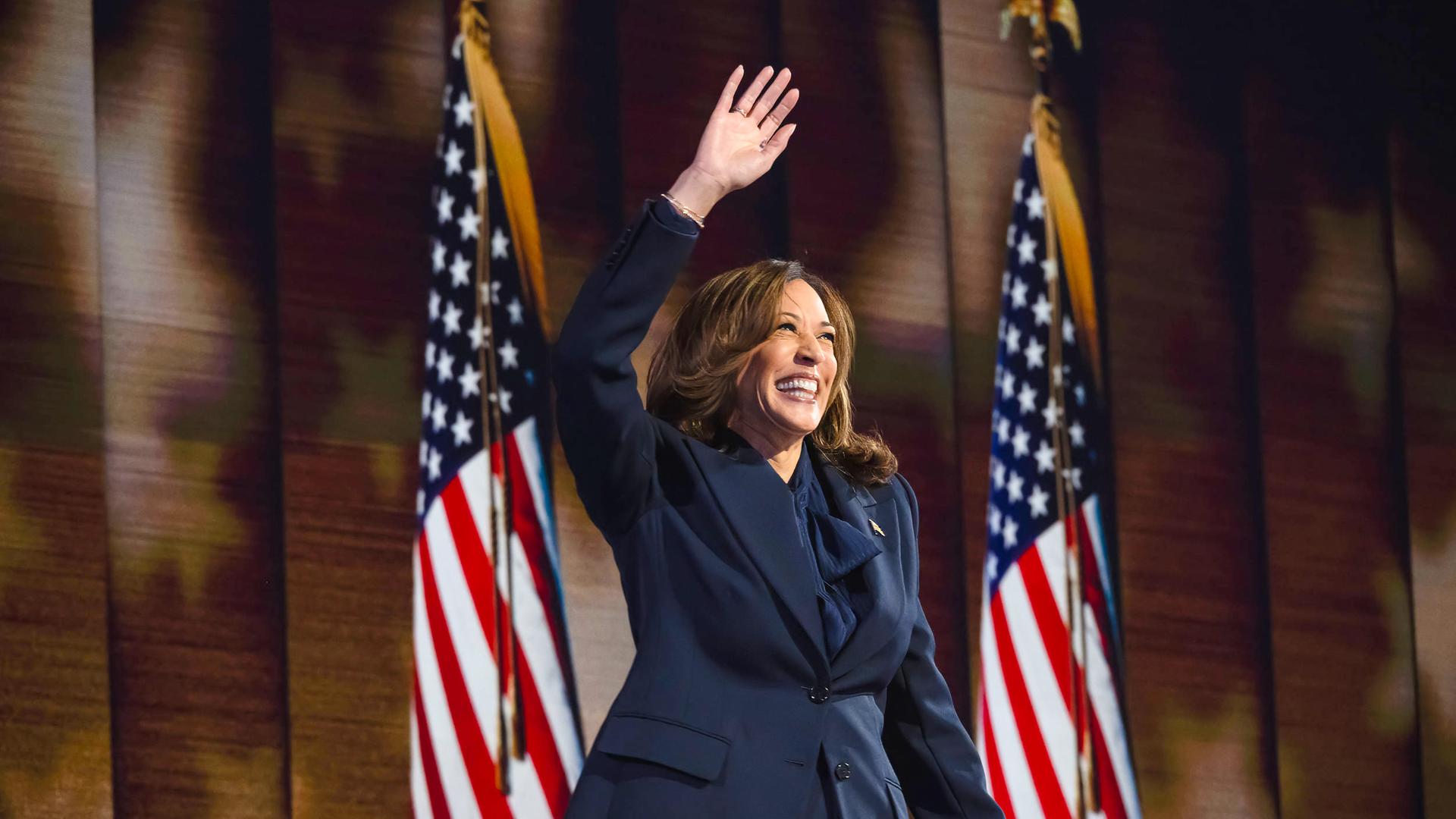 Eine Frau mittleren Alters mit langen braunen Haaren und gekleidet in einem Businessanzug steht zwischen zwei US-amerikanischen Flaggen und hebt lachend eine Hand hoch über den Kopf.