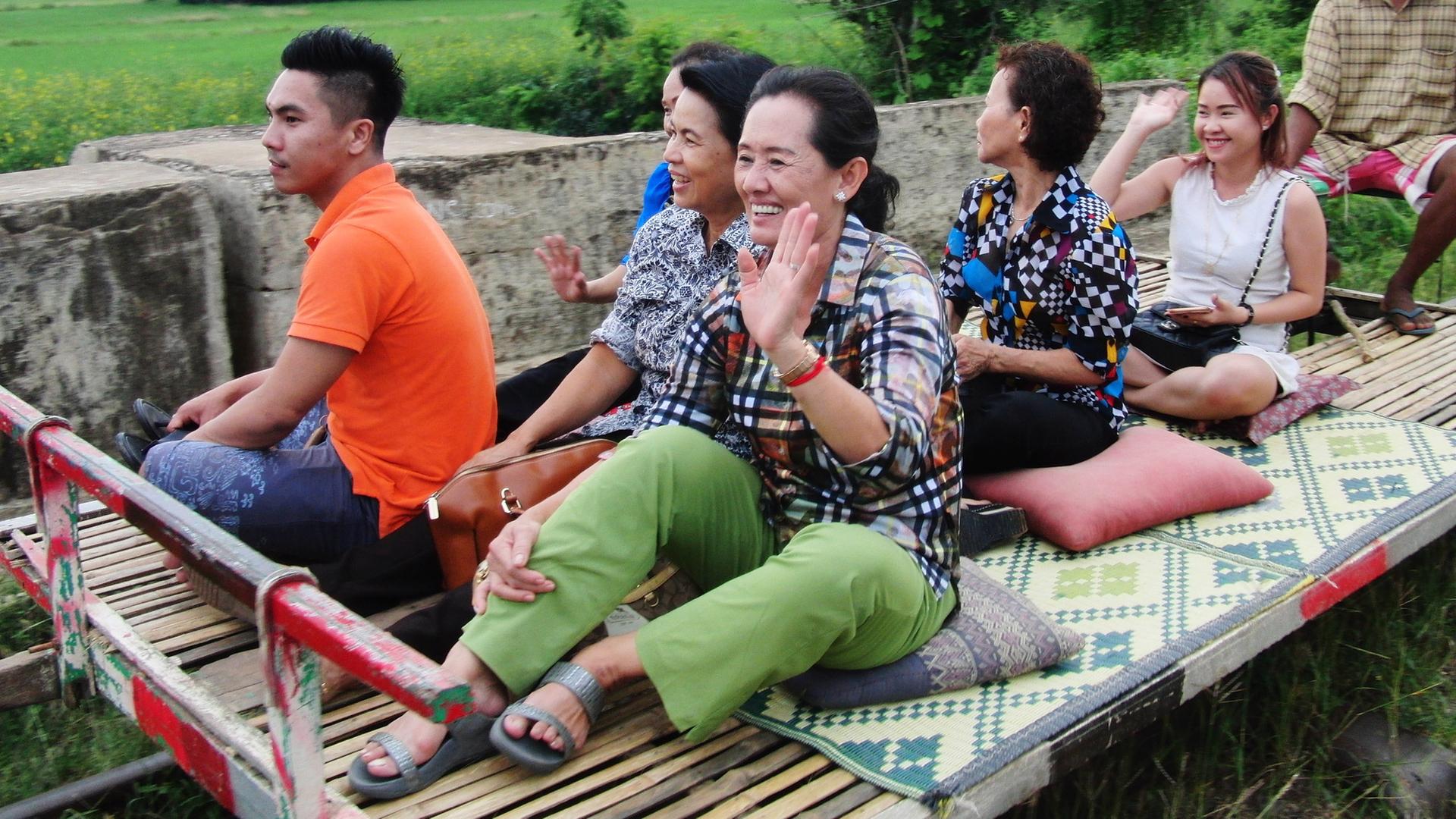Mehrere Menschen sitzen auf dem Wagen eines Bambuszugs und winken.