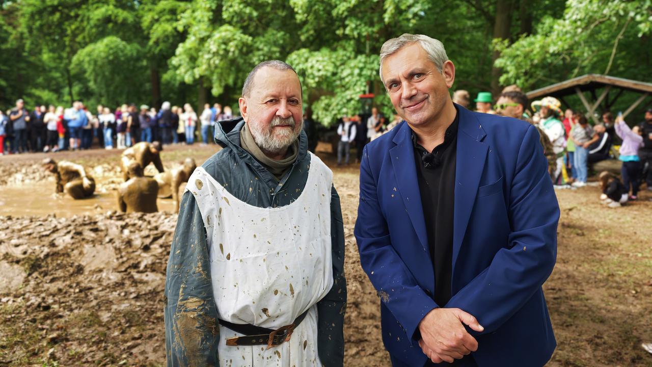 Vladimir Kaminer im blauen Anzug und eine Mann in einem weißen, schmutzigen Umhang, stehen lächelnd vor einer Gruppe von Menschen, die im Hintergrund zusehen. Einige Personen im Hintergrund spielen im Matsch. Im Freien, Bäume und Grün zu sehen.