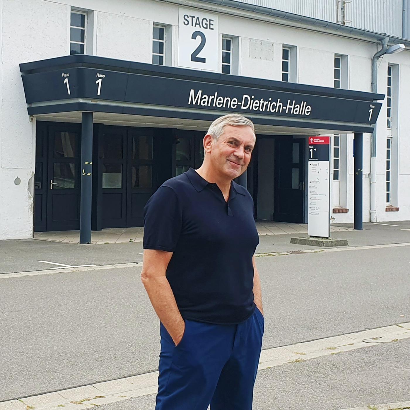 Wladimir Kaminer steht lässig mit blauem Poloshirt und blauer Hose, Hände in den Hosentaschen vor einer Hallenfront