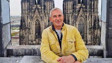 Ein Mann in gelber Lederjacke lehnt an einer steinernen Brüstung vor den Türmen des Kölner Doms; Blick über die Stadt Köln im Hintergrund. 