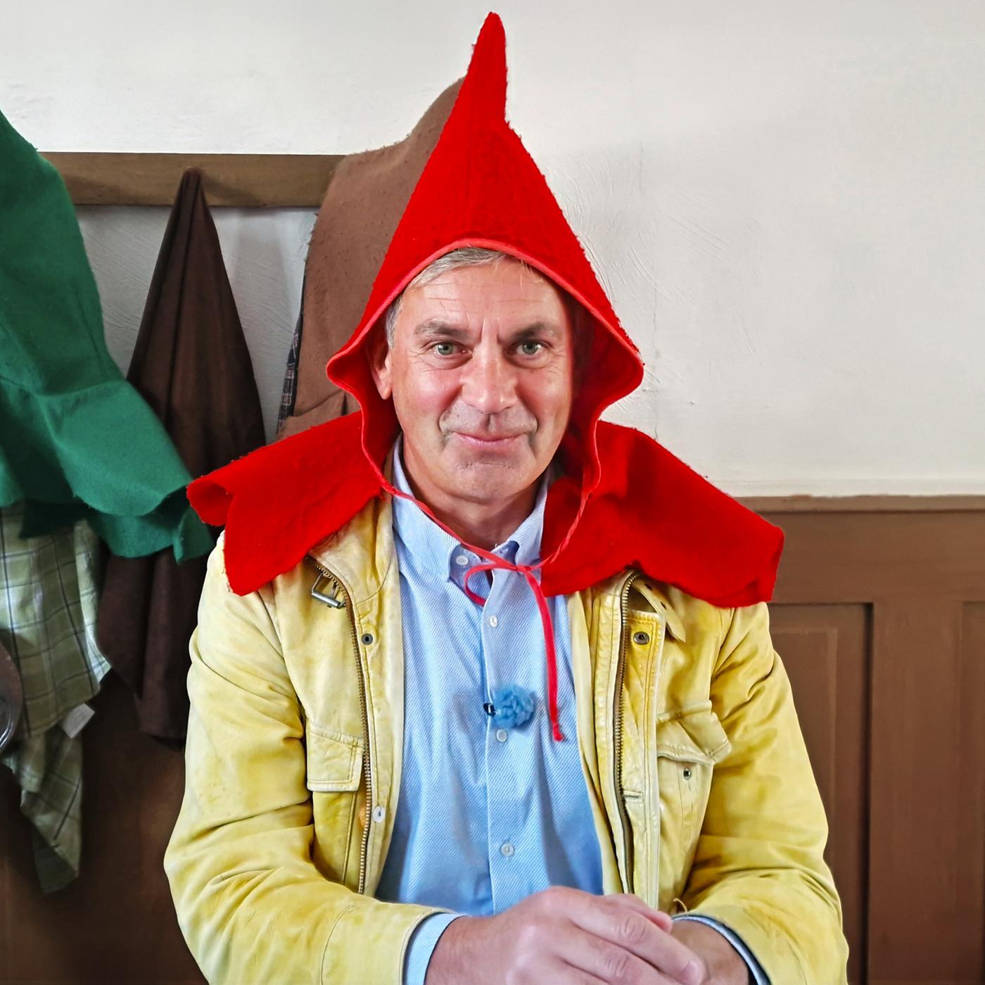 Wladimir Kaminer mit rotem Spitzhut und beiger Jacke an einem Tisch; im Hintergrund hängen bunte Hüte an der Wand -- Szene aus Kaminer Inside: Deutsche Märchenstraße.
