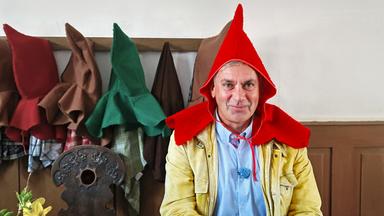 Wladimir Kaminer mit rotem Spitzhut und beiger Jacke an einem Tisch; im Hintergrund hängen bunte Hüte an der Wand -- Szene aus Kaminer Inside: Deutsche Märchenstraße.