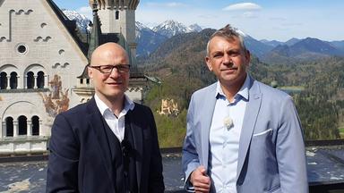 Kaminer und der Schlossherr von Neuschwanstein stehen nebeneinander auf einer Terrasse von Neuschwanstein, hinter ihnen Berge und Wälder