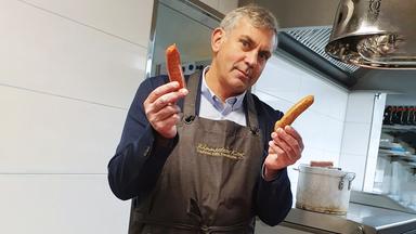 Wladimir Kaminer hält zwei Würstchen in der Hand. Vor ihm eine Schale mit Wurstwaren
