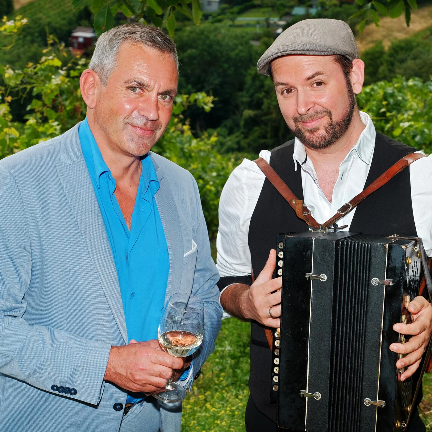 Zwei Männer stehen in einem Weinberg, einer hält ein Glas Wein, der andere ein Akkordeon.