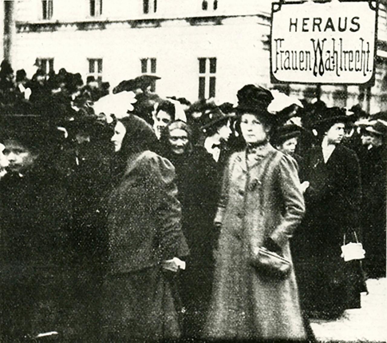 Das Bild zeigt eine historische Versammlung von Frauen, die für das Frauenwahlrecht kämpfen. Im Vordergrund sind mehrere Frauen zu sehen, die in Zeitkleidung aus der frühen 1900er Jahre gekleidet sind. Einige tragen Hüte und Mäntel, die typisch für diese Zeit sind. Hinter ihnen ist eine größere Gruppe von Frauen und Männern zu erkennen, die ebenfalls an der Versammlung teilnehmen. Es gibt ein Schild im Hintergrund, auf dem „HER AUS Frauen Wahlrecht“ steht, was auf die Forderungen der Frauenbewegung hinweist. Die Szene vermittelt eine Atmosphäre des Engagements und des Kampfes für die politischen Rechte der Frauen.   Der Hintergrund zeigt ein Gebäude, das in einem historischen Stil erbaut ist, was auf eine städtische Umgebung hinweist. Insgesamt strahlt das Bild den Geist einer Bewegung aus, die für soziale Gerechtigkeit und Gleichstellung eintritt.