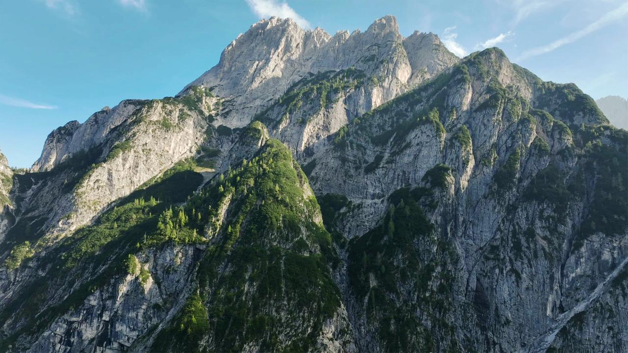Das Bild zeigt die Karnischen Alpen mit steilen, aufragenden Felsformationen und einer Vegetation, die sich an den Berghängen anpasst. Der Himmel ist blau mit einigen Wolken.