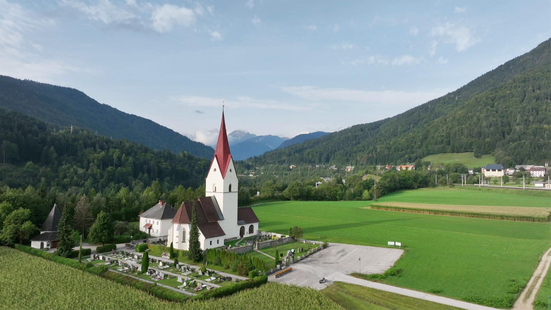 Das Bild zeigt eine Kirche mit rotem spitzen Turm, umgeben von grünen Wiesen und Bäumen. Im Hintergrund sind sanfte Hügel und Berge sichtbar. Ein Friedhof liegt neben der Kirche, sowie eine gepflasterte Fläche.