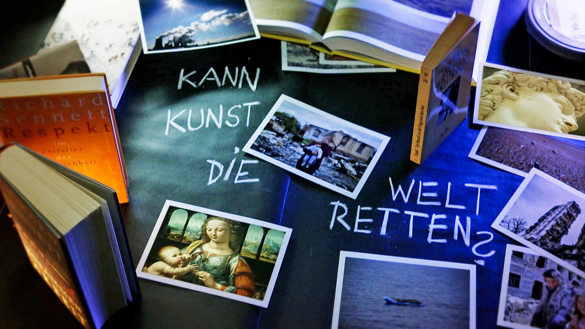 Auf einem Tisch sind viele Fotos verteilt, dazwischen steht mit Kreide geschrieben: Kann Kunst die Welt retten?