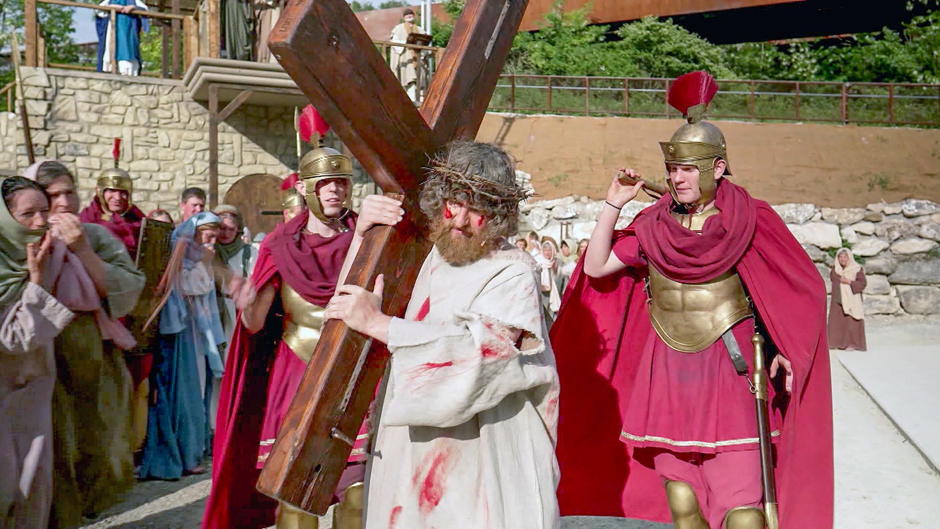 Das Bild zeigt eine Szene aus den Passionsspielen im Römersteinbruch von St. Margarethen. Eine Figur trägt ein Kreuz, umgeben von darstellenden römischen Soldaten und Zuschauern.