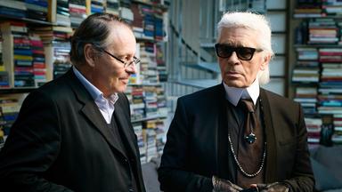 Karl Lagerfeld und Gero von Boehm stehen in einem Raum voller Bücher. Lagerfeld trägt ein schwarzes Outfit mit Krawatte, Sonnenbrille, schwarzen Lederhandschuhen und einer langen Perlenkette. Sein weißes Haar ist streng nach hinten gekämmt. Gero von Boehm, neben ihm, trägt einen dunklen Anzug mit Brille und scheint ihm zuzuhören. Im Hintergrund sind hohe Regale voller bunter Bücher sowie eine Couch mit Kissen zu sehen.